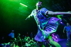 En dansare i färgstarka kläder snurrar på scenen med publik i bakgrunden, i en teatersal med dämpad belysning.