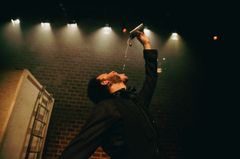 En skådespelare på scenen, klädd i mörka kläder, lutar huvudet bakåt och häller vätska från en plunta