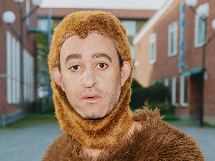 En person klädd i en apdräkt, med ett ansikte i bildens fokus. Byggnader syns i bakgrunden.