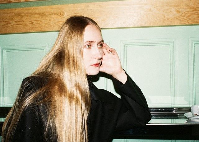 Kvinna med långt blont hår sitter vid ett bord och tittar framåt.