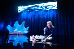 Ann-Sofie sitter på en mörk scen med blå belysning. I bakgrunden finns en stor, vit snäcka.