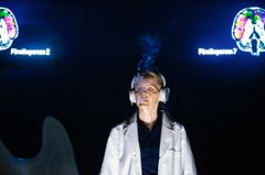 Ann-Sofie Rase står på scen med hörlurar och en vit labbrock på sig. Hon blundar och blåser ut rök ur sin mun.