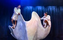 Emil Hedayat och Angelika Prick i vita kläder poserar på en stor, abstrakt scenografi föreställande en öppen snäcka.