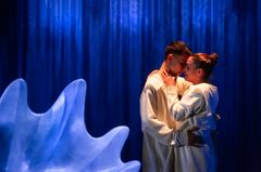 Två personer i vita kläder står nära varandra på en scen med blått ljussken.