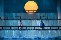 Två personer står på en dramatisk scen med en stor gul cirkel i bakgrunden.