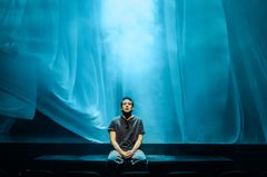 En person sitter på en scen med mjukt blått ljus och draperier i bakgrunden.