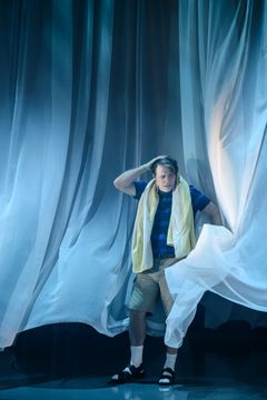 En man i shorts och sandaler med en handduk runt nacken, omgiven av svepande vita draperier.