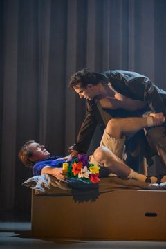 Två personer på scen, den ena ligger på en säng med en färgglad bukett blommor, den andra står över och lutar sig mot den första.