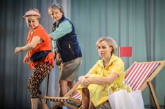 Tre skådespelare framför en scen med strandtema, en av dem sitter på en solstol.