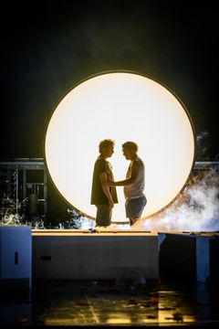 Två skådespelare på en scen med en stor ljuscirkel i bakgrunden.