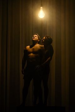 Två personer står nära varandra på en scen under en enkel glödlampa.