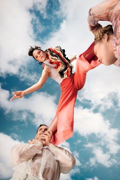 Två cirkusartister som lyfter upp en tredje artist i färgglada kläder mot en blå himmel med några moln.