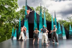 Grupp av cirkusartister på en utomhusscen framför en backdrop med blå spetsiga former.