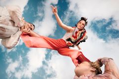 Två cirkusartister som lyfter upp en tredje artist i färgglada kläder mot en klarblå himmel med några moln.