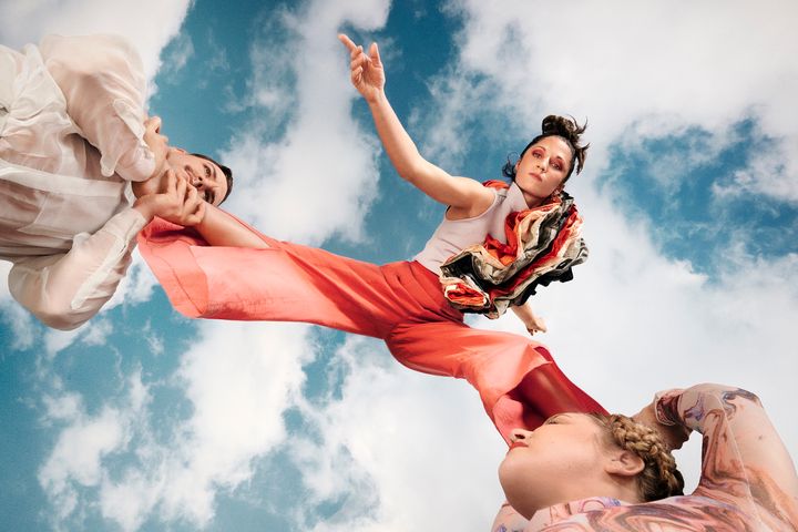 Två cirkusartister som lyfter upp en tredje artist i färgglada kläder mot en klarblå himmel med några moln.