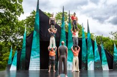 Grupp av sex personer utför en cirkusföreställning utomhus, omgiven av en kreativ, spetsig scenografi.