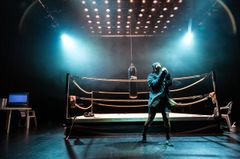 Man i boxningskläder tränar under en boxningssäck i en upplyst ring med en dator på ett bord i bakgrunden.