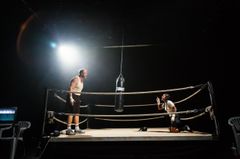 Två personer i en boxningsring; en står upp och den andra sitter på knä. En boxningssäck hänger mellan dem.