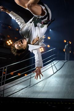 En person i en vit kostym gör en akrobatisk rörelse i luften på en scen med stark belysning.