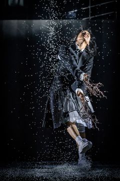 Bild av en person i en dramatisk pose på scen, omgiven av fallande vita partiklar.