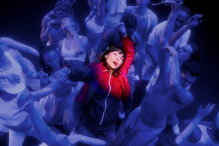 En kvinna i en röd jacka i centrum, upplyst i en folkmassa som dansar runt henne. Scenen är upplyst i blått ljus.