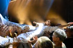 Skådespelare i en vit kostym blir upplyft av publiken under en teaterföreställning.