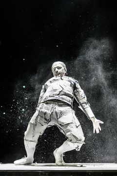 En person i en dramatisk pose, klädd i vit, konstnärligt rivet kostym, med vit färg och pulver runt omkring.