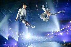 Två artister utför akrobatiska hopp på en teaterscen med ljusuppsättning i bakgrunden.