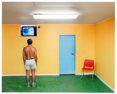 Man utan tröja står i ett rum med gula väggar och grön matta, tittar på en liten TV högt upp på väggen. En röd stol står bredvid en blå dörr.