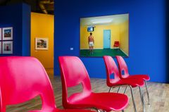 En konstutställning med röda stolar i förgrunden och en tavla på en vägg med en man i ett gult rum.