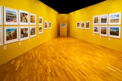 Alt text: Bild av en konstutställning med färgglada fotografier hängda på gula väggar i ett galleri.