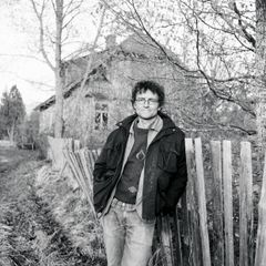 En person står framför ett trästaket med ett gammalt hus i bakgrunden. Det är en vinter- eller höstdag.