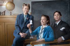 Tre personer i ett klassrum från tidigt 1900-tal som är klädda i äldre stil. En kvinna sitter lutad mot skolbänken, framför henne sitter en annan kvinna som håller en papperssvala och en man i kostym. I bakgrunden syns en jordglob och en griffeltavla.