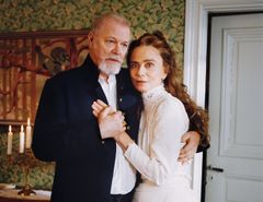 Bilden visar Peter Andersson och Lena Olin i periodkläder, de håller varandra i handen i en inomhusmiljö med blommig tapet och en dörr i bakgrunden.