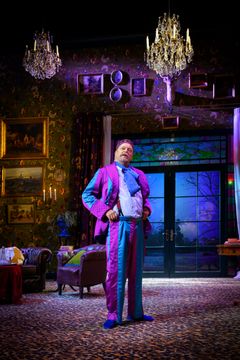 Peter Andersson står på scen i en färgglad kostym, omgiven av inredning som inkluderar ljuskronor och målningar.