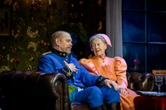 Peter Andersson och Lena-Pia Bernhardsson i kostymer sitter på en soffa, i en scen från en teaterföreställning i en hemtrevlig miljö.