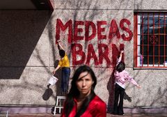 Två personer målar texten "Medeas Barn" i rött på en vägg. En person står i förgrunden.