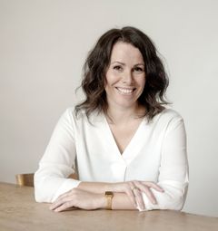 Kvinna med mörkt hår och vit blus sitter vid ett bord och ler.