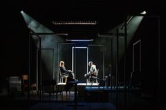 En dunkel teaterscen med två personer, en man och en kvinna, som sitter vända mot varandra.