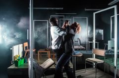 Två personer bråkar på en scen med minimalistisk inredning.