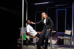 En person i kostym står och sträcker sig mot en sittande person på en teaterscen.