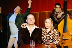 Två personer sitter vid ett bord, en står med en kontrabas och en person som dansar.