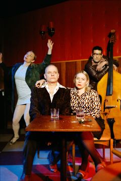 Två personer sitter vid ett bord, en står med en kontrabas och en person som dansar.