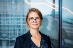 Porträtt av en person med glasögon och mörk kavaj framför ett fönster med stadsbakgrund.