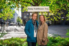 Två personer står framför en skylt som säger "Benny Fredrikssons Torg" i en solig stadsmiljö.