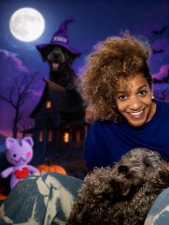 Kvinna ler med en hund i famnen framför ett halloween-tema med spökhus, måne och en leksak.