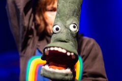 En person håller upp en grön stövel med ögon och mun som del av en teaterföreställning.