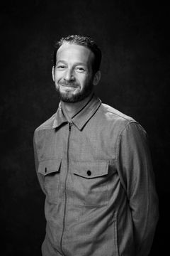 Jag vet inte vem det är, men här är en möjlig bildbeskrivning: En man med kort hår och skägg, står klädd i en jacka med fickor fram, framför en mörk bakgrund.