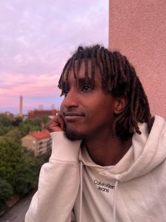 En person med lockigt hår blickar ut över ett stadslandskap i solnedgången.