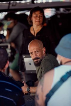 Alt-text: Man med skägg sitter vid ett bord och lyssnar, omgiven av andra personer.